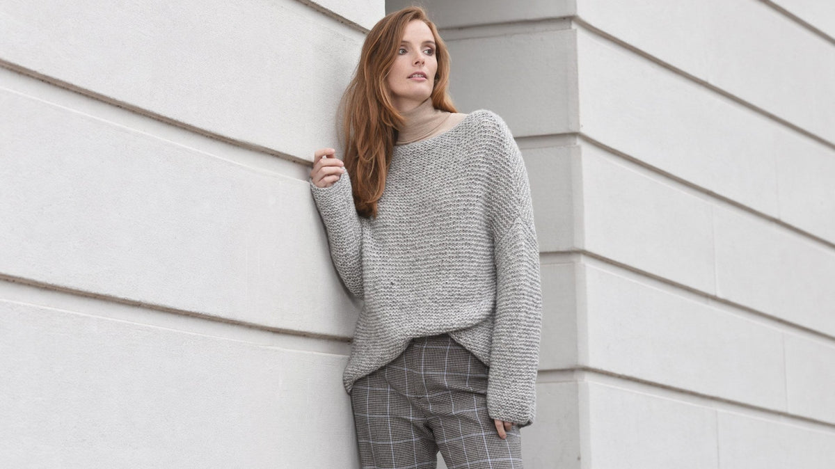 Model trägt Pullover Nora in kraus rechts vor einer hellen Steinwand. Originalfoto, Hintergrund seitlich mit KI erweitert; Person, Pullover und Farben unverändert.