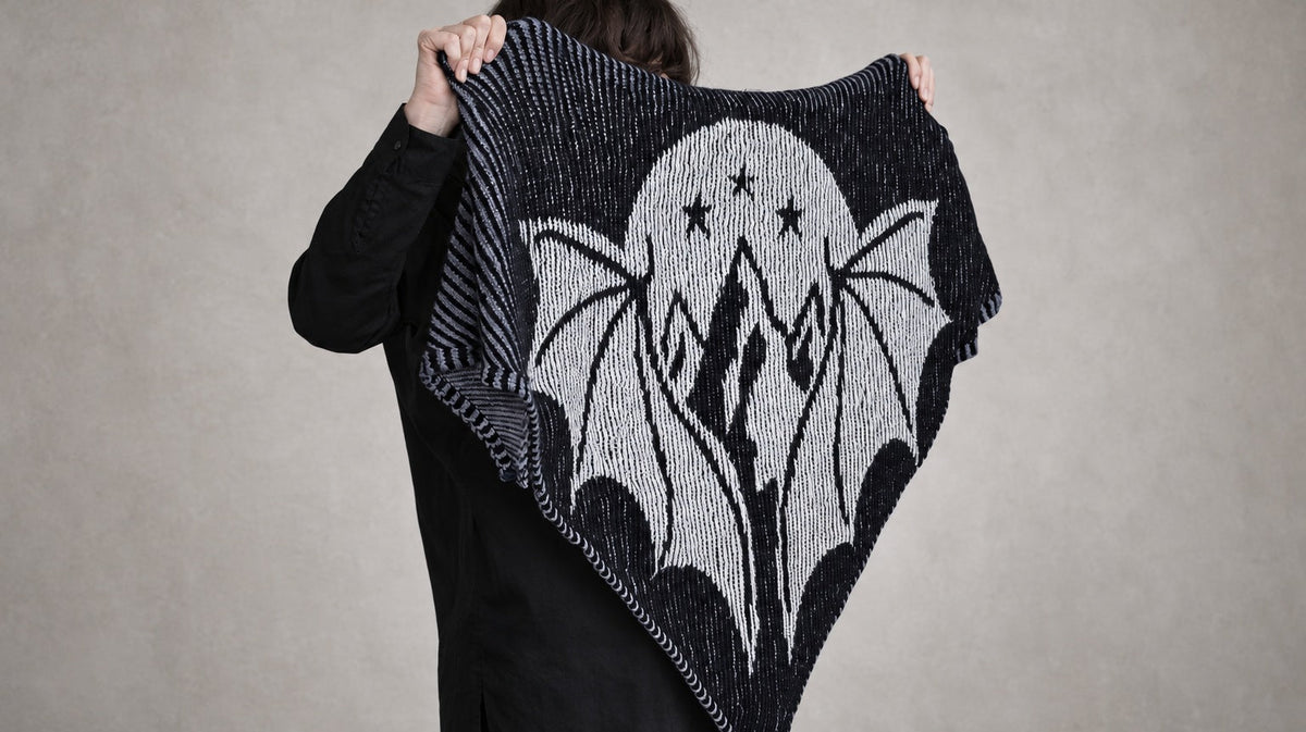 Claudia zeigt das schwarz-weiße Schattenstrick-Tuch Starborn mit Flügeln, Bergen, Mond und Sternen. Originalfoto, Wandhintergrund für das Querformat mit KI erweitert.