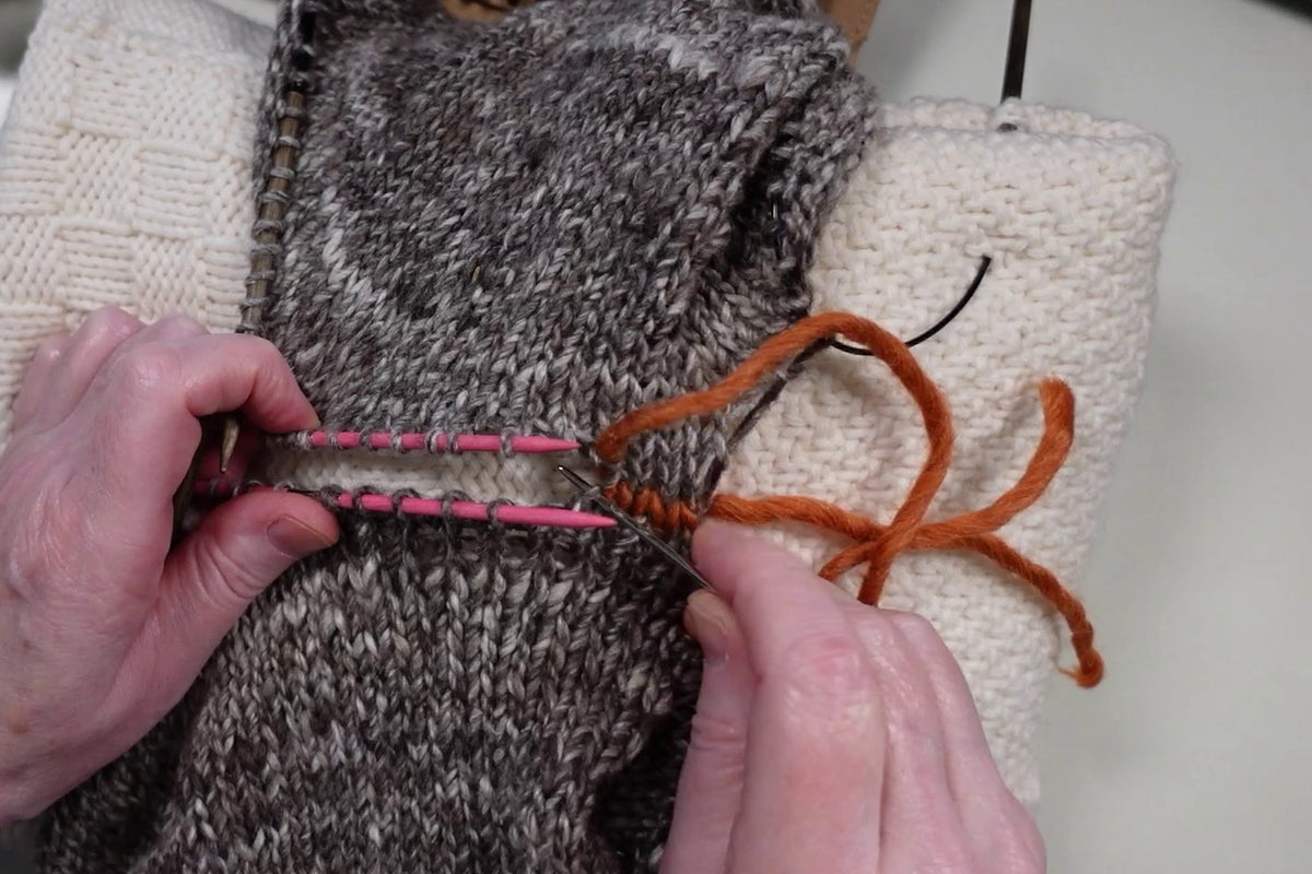 Orangefarbener Maschenstich verbindet die offenen Maschen zweier grauer Strickteile an pinken Nadeln.