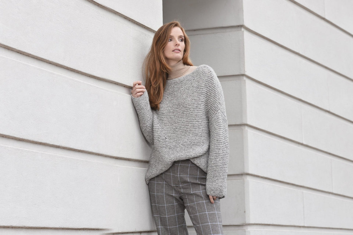 Model trägt Pullover Nora in kraus rechts vor einer hellen Steinwand. Originalfoto, Hintergrund seitlich mit KI erweitert; Person, Pullover und Farben unverändert.