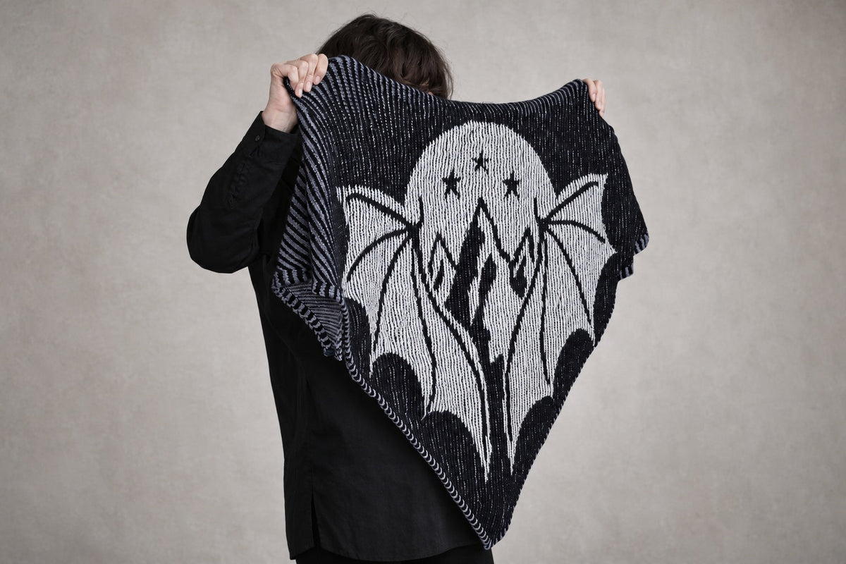 Claudia zeigt das schwarz-weiße Schattenstrick-Tuch Starborn mit Flügeln, Bergen, Mond und Sternen. Originalfoto, Wandhintergrund für das Querformat mit KI erweitert.
