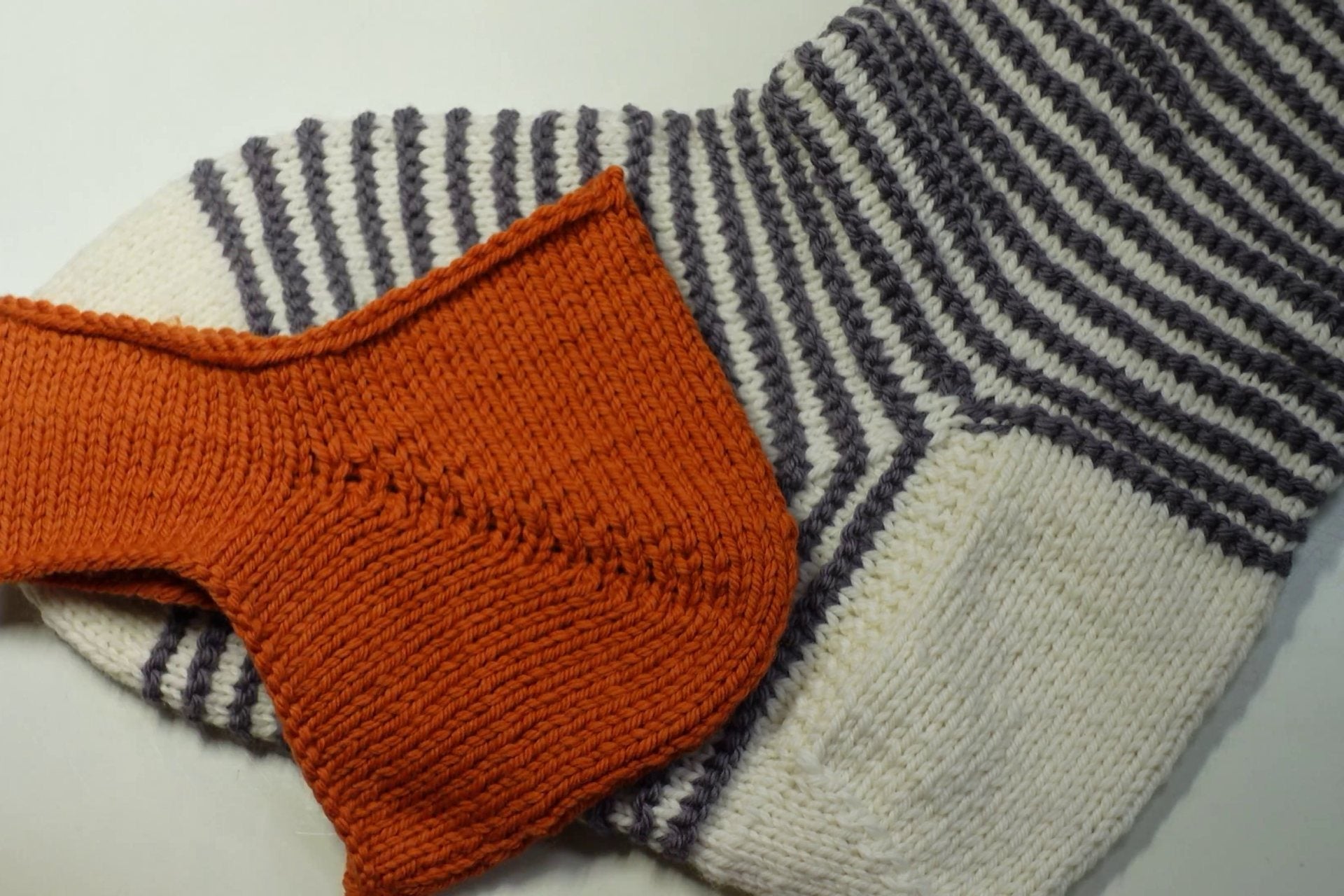 Gestrickte Bumerangferse in Orange vor einer geringelten Socke
