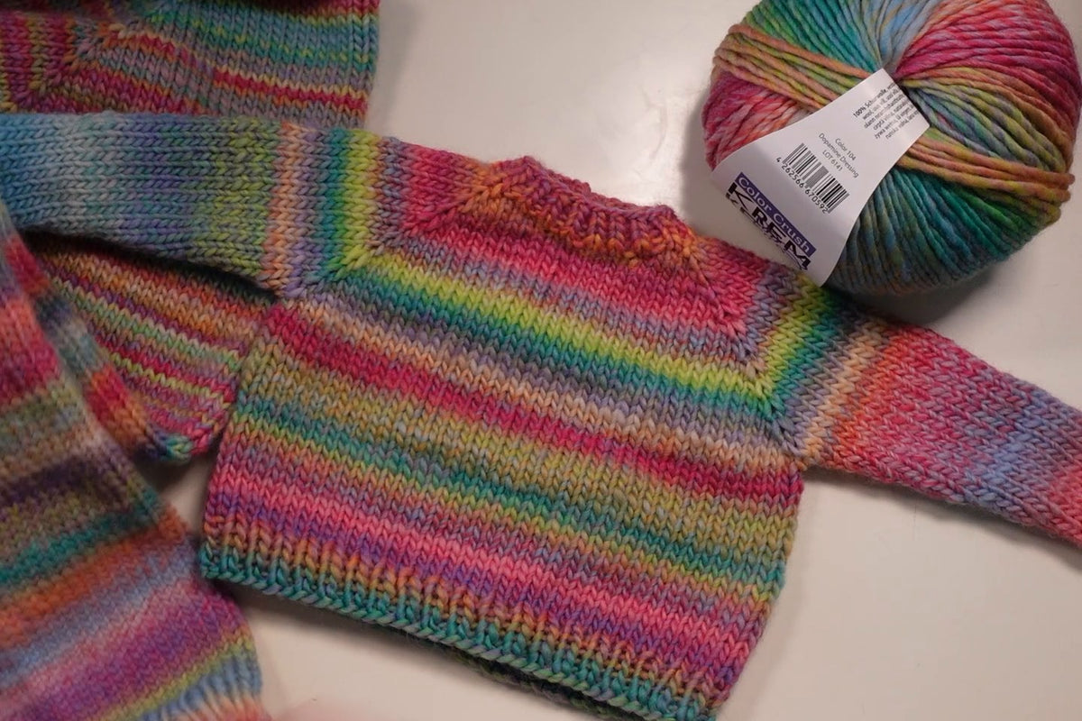 Kleiner bunt geringelter Raglanpullover flach auf dem Tisch, daneben ein Knäuel Color Crush von Kremke